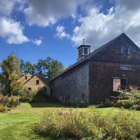 A large, weathered wooden barn with a cupola stands beside a smaller farmhouse, surrounded by a grassy lawn, shrubs, and autumn trees under a partly cloudy blue sky.
