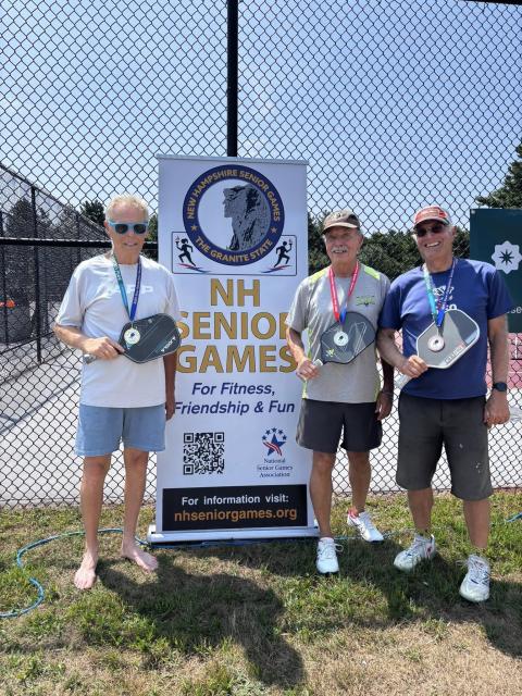 Three men standing in front of a banner for NH Senior Games, holding pickleball paddles, indicating participation in a senior fitness event.