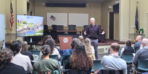 A man giving a presentation to an audience seated in a room with American flags and a podium. A large screen displays a slide titled "District 6 Economic Development" with a cityscape background, indicating a community or economic development meeting.