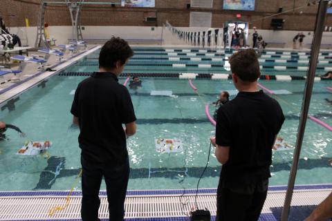 Two youth participate in the Seacoast SeaPerch Challenge. They are both in black shirts, their backs to the camera, looking into an indoor pool at their underwater remote-controlled vehicle.