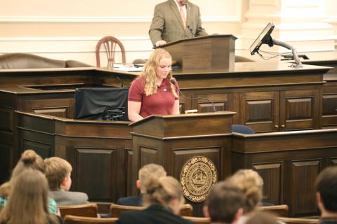 A young woman with blonde hair and wearing a maroon shirt stands at a podium in the NH Statehouse. She is speaking into a microphone. Audience members can be seen in the foreground of the photo.
