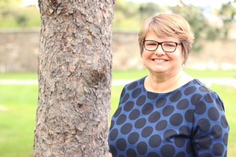 Extension specialist Molly Donovan stands next to a tree. She is seen from the chest up. She is waering a blue shirt with dark blue polka dots and glasses. She is smiling at the camera.