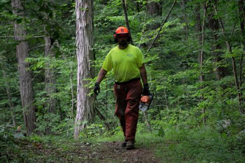 a man in a brightly colored shirt wearing a hardhat and chaps and carrying a chainsaw in a forest setting
