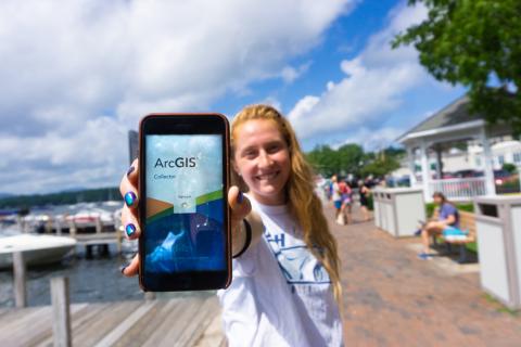 Community and Economic Development intern Hannah Stewart holds a phone running the ArcGIS Collector app. She is holding the phone to the camera and smiling. Behind her is a harbor full of boats. People walk behind her along a sidewalk.