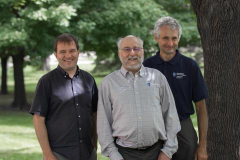 Three men, one in a blue collard shirt on the left, one in a light blue shirt and with a beard and glasses and the center, and one on the right in a blue polo shirt and standing next to a tree, smile and face the camera.