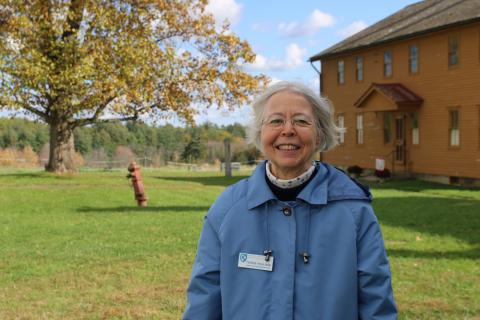 An older woman in a blue jacket and waering glasses smiles at the camera. She is standing in front of a large historical home in Portsmouth.