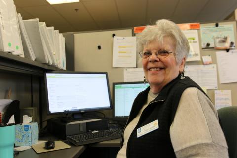 An older woman with white hair and glasses sits at a desk. she is turned to face the camera. she is wearing a tan shirt and a black fleece vest. she is smiling.
