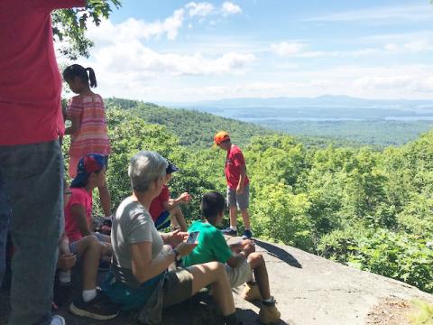 Adults and children gatherd on cliff looking over large amount of woods