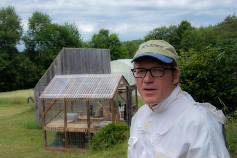 A man in white beekeeping gear and a tan hat looks at the camera. He is smiling. Behind him are various farm structures.