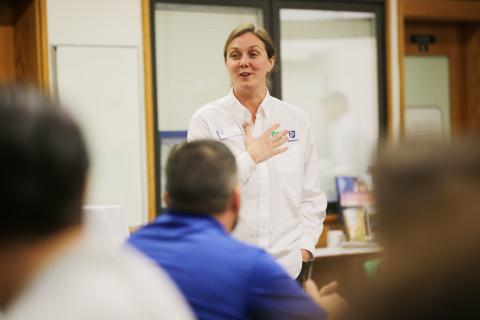 A woman in a white shirt addresses an audience in a classroom. The audience is visible in the foreground of the photo. She is smiling and holding her hand to her chest.