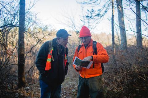 A photo of two men in the woods. The man on the right is wearing an orange jacket and hat and is pointing to an informational handout; the man on the left is wearing a dark jacket and hat and an orange vest and is looking at the map the other man is holding.