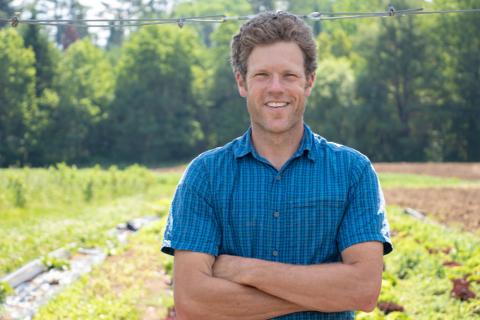 A man in a short-sleeved blue button-down shirt stands facing the camera. His arms are crossed and he is smiling. Behind him is a field of crops on a farm.