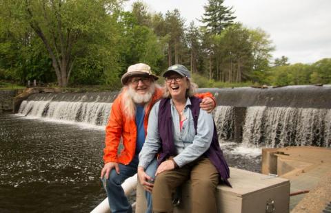 A man and a woman sit on a wooden box in front of a waterfall at a dam. They are both smiling. They are older; the man has long white hair and a white beard and is wearing an orange jacket and glasses. The woman has long gray hair and is wearing glasses and a blue shirt and purple vest. The man has his arm around the woman.