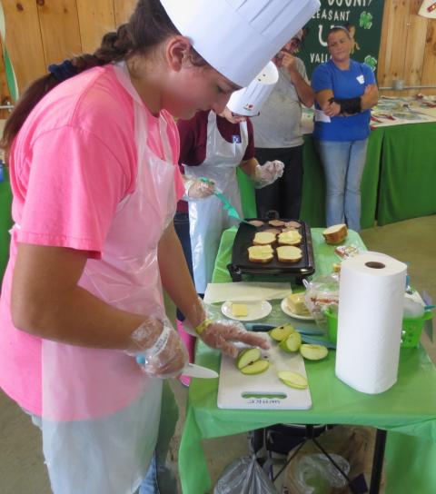 4-H members make grilled cheese sandwiches during a competition. In the photo, two members are wearing chefs hats and aprons and looking down a griddle. Behind them, two people look on.
