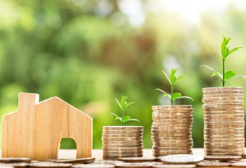 Stacks of coins with plants appearing to grow from them, next to a wooden model house