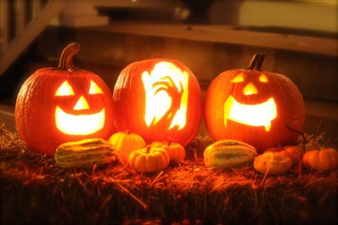 Three jack-o-lanterns on a bale of hay. The jack-o-lanterns have faces carved into them and they are lit by candles. Smaller decorative gourds surround them.