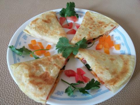 A quesadilla cut into four slices and arranged in a cross shape on a plate. Chopped orange and red peppers and pieces of parsley are between the slices. The plate is white with a blue pattern.
