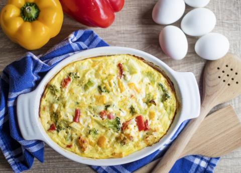 A photo of a quiche in a white serving dish. A blue and white towel surrounds the dish; next to the dish are, on the left, a yellow and a red pepper, and on the right, four eggs.