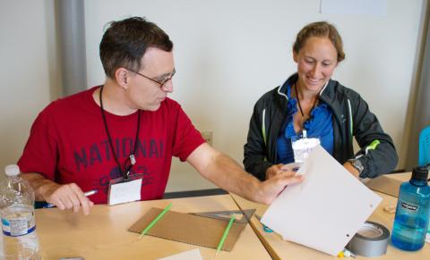Two teachers sit together at a table. They are working on a science project with materials spread out in front of them. On the left is a man in glasses and a red t-shirt; on the right is a woman in a blue shirt and a dark green jacket.