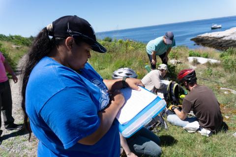A woman in a blue shirt and cap is writin gsomething on a clipboard. Behind her, five volunteers are engaged in activities on a rocky, grassy hillside.