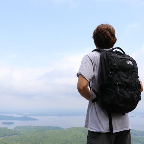 Hiker wearing backpack, looking out towards lake atop a mountain